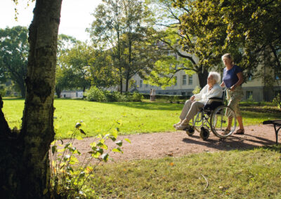 Eine Frau schiebt eine Seniorin im Rollstuhl im Park Akazienhof