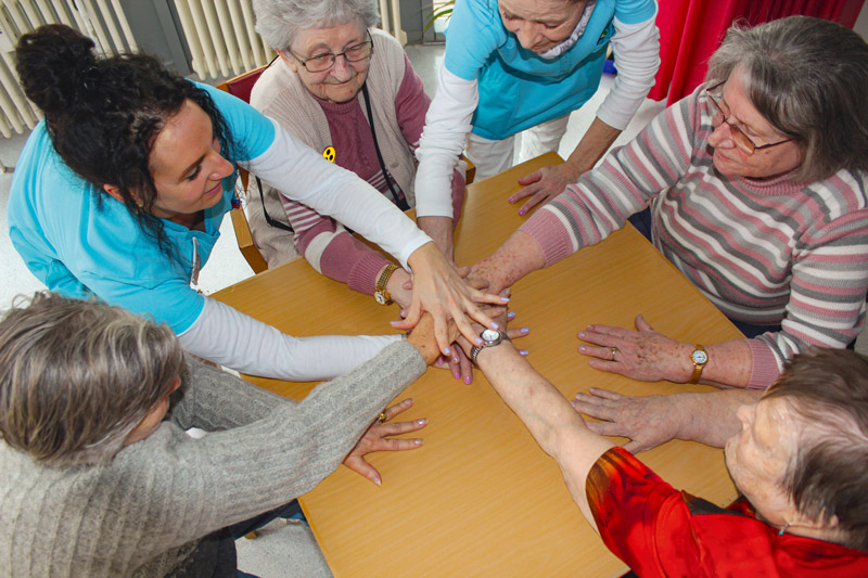 Tagespflege in Halle (Saale). © 2023 Paul-Riebeck-Stiftung Mehrere Seniorinnen und Pflegekräfte spielen ein Spiel am Tisch mit Händeauflegen