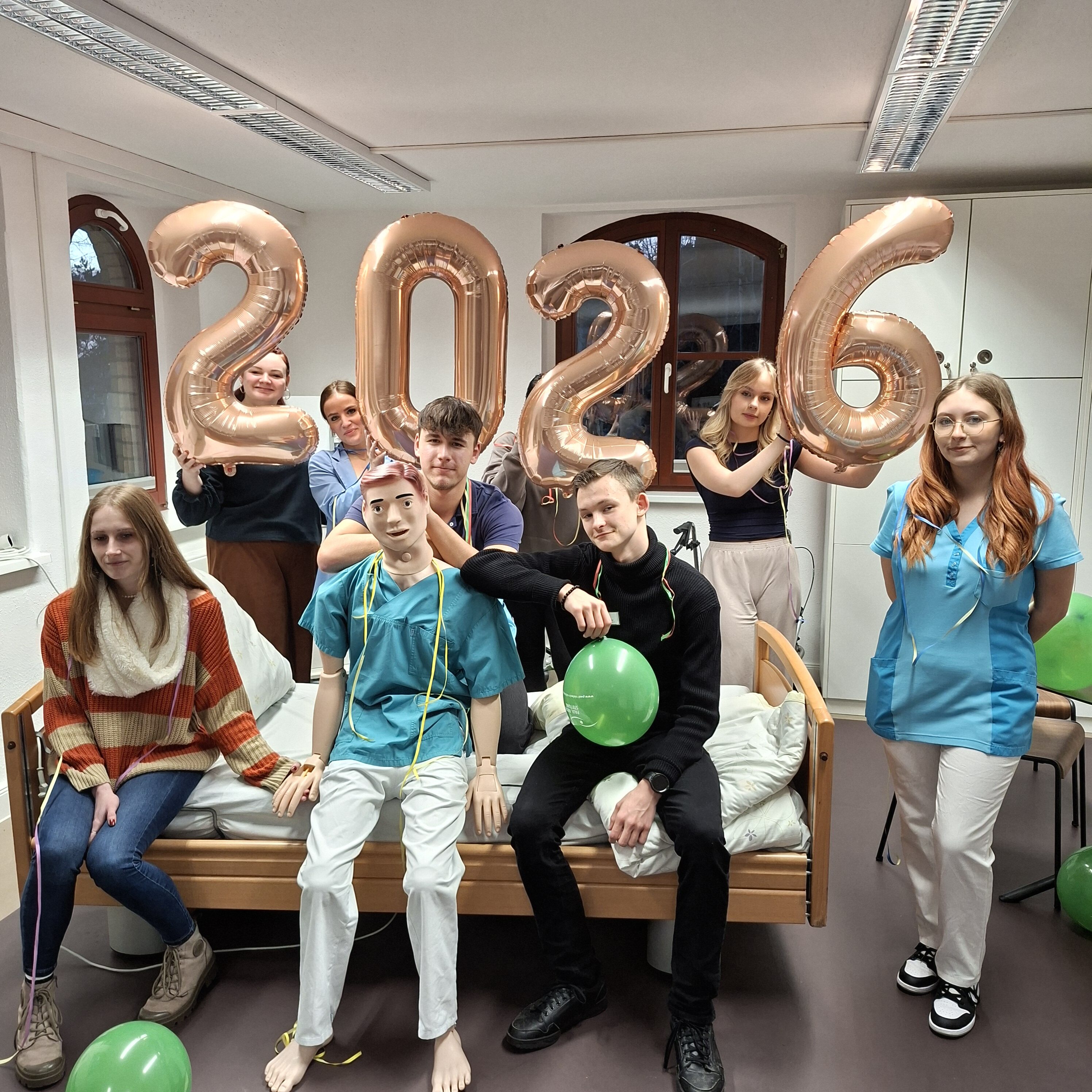 Ein gutes Neues Jahr von unseren Azubis und Pflegepuppe Helmut! © Paul-Riebeck-Stiftung Acht Auszubildende der Paul-Riebeck-Stiftung im Praxis-Raum mit Trainingsraum mit Pflegepuppe und Luftballons in Form einer 2026